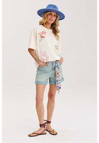 Krémové tričko s farebnými grafikami, skombinované s svetlo modrými denimovými šortkami. Modrý široký klobúk, členený šál zviazaný v páse, ploché sandály.