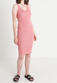 Abito rosa lavorato a maglia con scollatura a V e taglio aderente, caratterizzato da spalline larghe, orlo a ginocchio e tessuto texturizzato. Sandali neri con fibbie.