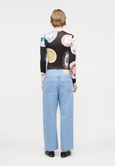 Άτομο με κοντά ξανθά μαλλιά φοράει τζιν ψηλής μέσης ανοιχτού μπλε χρώματος και μακρυμάνικη μπλούζα με πολύχρωμα κυκλικά σχέδια, στέκεται γυρισμένο με την πλάτη.