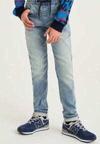 Bambino che indossa jeans azzurri chiari, sneakers New Balance blu navy e bianche, e una maglietta a maniche lunghe dai colori vivaci blu, rosso e nero.