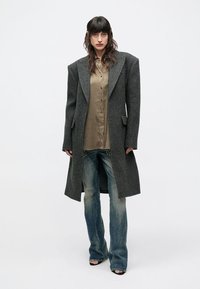 Sivý vlnený kabát so širokými klopami, veľkými vreckami a dlhým strihom, doplnený béžovou košeľou na gombíky a svetlomodrými džínsami.