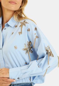 Lichtblauwe button-up shirt met florale geborduurde ontwerpen in goud, met een relaxed fit en opgerolde mouwen.