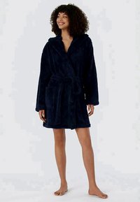 Woman standing barefoot wearing a dark blue hooded bathrobe tied at the waist, smiling and looking to her left.