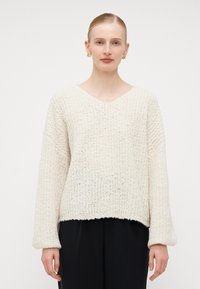 Megzta suknelė off-white spalvos, su V formos iškirpte, nukritusiais pečiais ir ilgomis rankovėmis. Audinys su tekstūra, atrodantis minkštas ir storas.