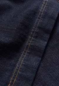 Mörk denimtyg med en jämn textur, med kontrasterande orange sömnad längs en söm, som visar på fin väv och djup.