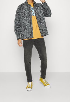 Chaqueta gris con patrón y diseños de "Santa Cruz", camiseta amarilla, vaqueros oscuros y zapatillas amarillas. La chaqueta tiene un corte relajado y botones.