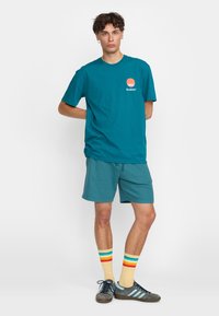 Een teal katoenen T-shirt met een oranje cirkelvormige graphic en het 'Revolution'-logo. Gecombineerd met bijpassende shorts; het model draagt meerkleurige gestreepte sokken.