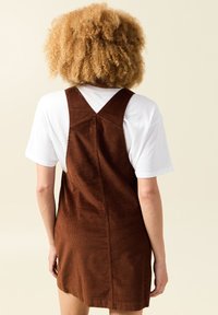 Braunes Cord-Overall-Kleid mit taillierter Silhouette und überkreuzten Trägern, getragen über einem weißen T-Shirt. Glatte Textur, knielang.