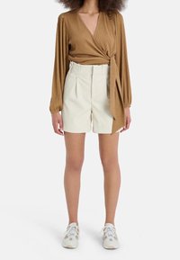 Top avvolgente marrone a costine con maniche lunghe, abbinato a pantaloni corti crema a vita alta. Il modello indossa sneaker bianche con accenti geometrici.