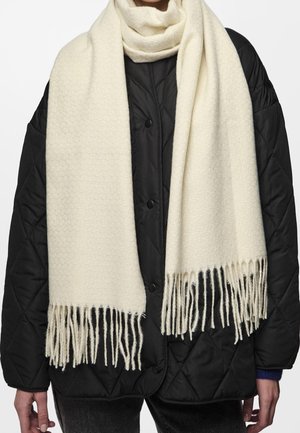 Écharpe en laine crème avec franges portée sur une veste matelassée noire à boutons-pression par une personne.