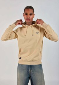 Homme ajustant la capuche d'un sweat à capuche beige avec poche avant et petit logo, portant un jean bleu sur un fond clair uni.