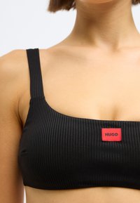 Črni rebrasti bralette z širokimi naramnicami in kvadratnim izrezom; na sprednji strani majhna rdeča oznaka »HUGO«. Gladek tekstura.