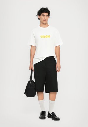 T-shirt bianco in cotone con design grafico giallo, pantaloni corti neri su misura, mocassini neri e calzini grigi. Modello che tiene una borsa nera.