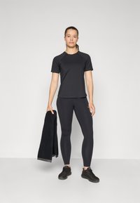 Svart atletisk t-shirt och leggings i ett glatt, stretchigt tyg, i kombination med svarta skor, hållande en mörk handduk.