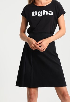 Vrouw met een zwart t-shirt met "tigha" in witte letters en een zwarte rok, staat met haar handen gevouwen voor zich.