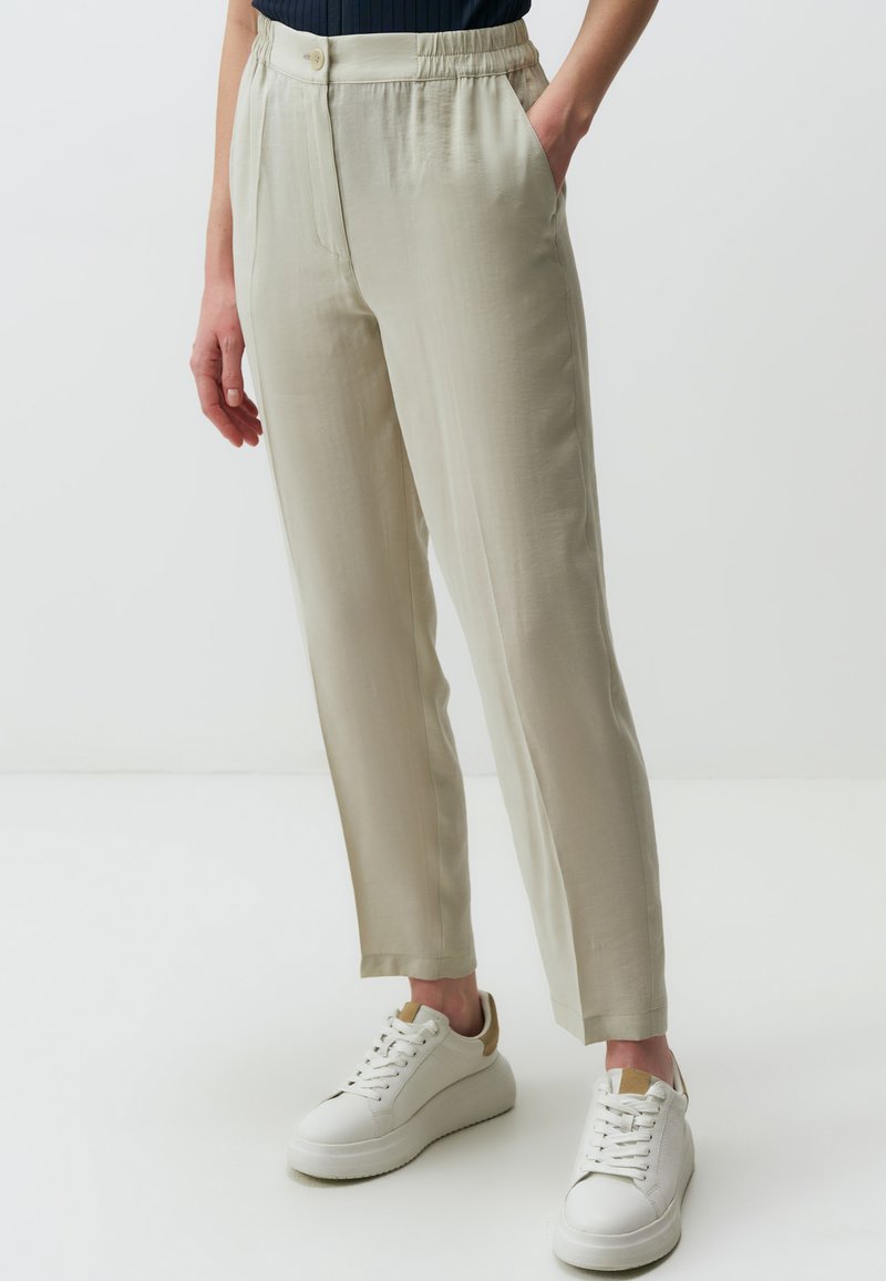 Pantaloni elasticizzati di colore beige chiaro con gamba dritta. Presentano una chiusura con bottone e tasche laterali. Indossati con sneakers bianche.