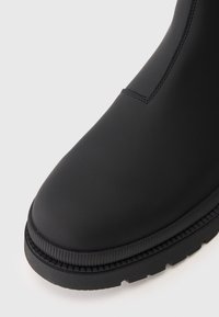 Bota de tornozelo preta em borracha com acabamento mate suave, bico arredondado e sola texturizada. Costura visível na parte superior.