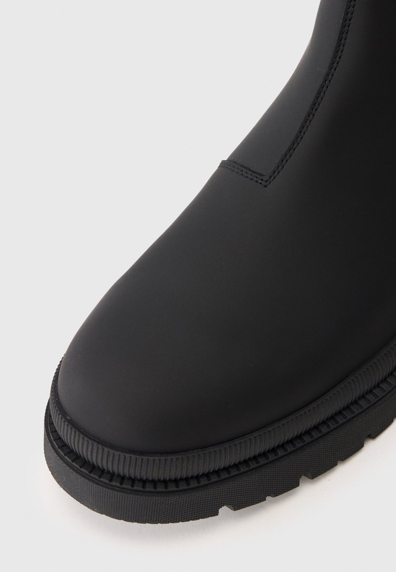 Zwarte rubberen enkelboot met een gladde matte afwerking, ronde neus en een gestructureerde zool. Naaddetailing zichtbaar op de bovenkant.