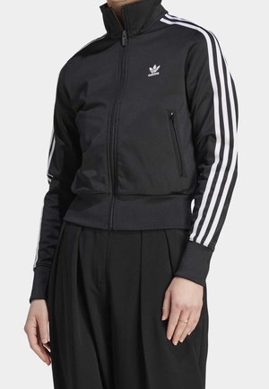 Schwarze Adidas Zip-up Jacke mit weißen Streifen an den Ärmeln und Logo auf der Brust, getragen mit lockeren schwarzen Hosen.