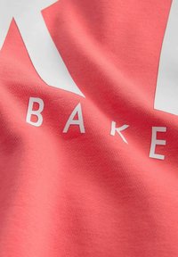 Koralļu kokvilnas audums ar gludu tekstūru, uz kura ir balta zvaigznes forma un vārds "BAKE" taupīgās burtiem visā dizainā.