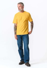 Gul bomulds-t-shirt med korte ærmer, parret med blå denim jeans. Emnet har tatoveringer og bærer brune ruskindsko.