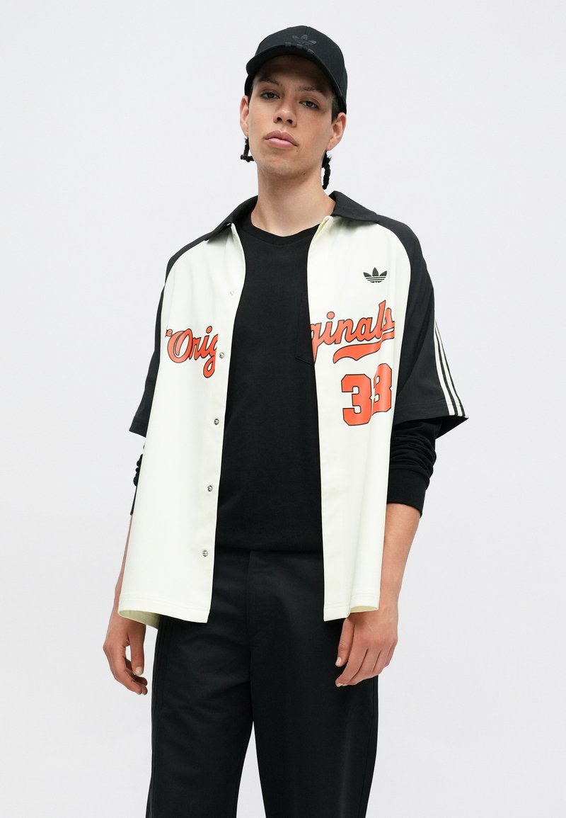 Witte en zwarte overhemd met knopen met "Originals 38" in oranje, gedragen met een zwart t-shirt en bijpassende pet, in combinatie met zwarte broek.