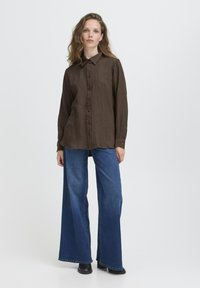 Frau mit langen,Welligen Haaren, die ein braunes Knopfshirt, weit geschnittene blaue Jeans und schwarze Schuhe trägt und vor einem schlichten weißen Hintergrund steht.
