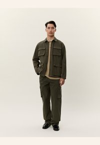 Giovane uomo in piedi, indossa una giacca verde oliva e pantaloni cargo con una camicia beige, scarpe nere, sfondo neutro.