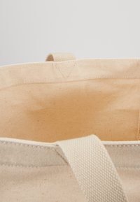 Bomullstoteväska i ljusbeige färg, med ett texturerat tyg. Inuti finns en slät foder utan ytterligare fickor eller avdelningar.