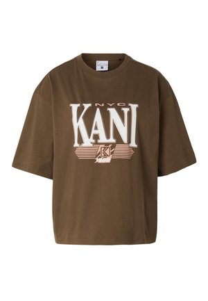 Femme blonde aux cheveux lâchés, portant un t-shirt KANI marron oversized et un jean bleu clair, debout avec une main dans la poche.