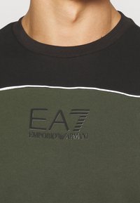 T-shirt corto verde e nero con collo rotondo. Presenta un logo EA7 Emporio Armani testurizzato e dettagli in bianco a contrasto.
