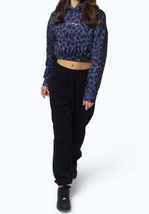 Kvinde iført en cropped hættetrøje med blå og sort leopardprint og løse sorte bukser, der går fremad mod en hvid baggrund.