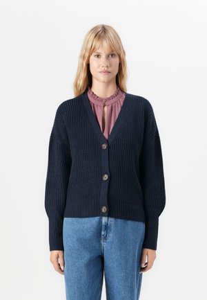 Femme portant un cardigan boutonné bleu marine sur un blouse mauve à volants et un jean bleu clair, sur un fond blanc.
