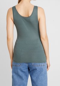 Canottiera a coste in tessuto verde scuro, con spalline larghe e orlo curvo; indossata con jeans in denim azzurro chiaro.