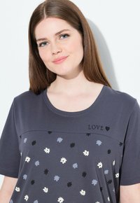 Žena s rovnými hnědými vlasy, která má na sobě tmavě šedý květinový top s výšivkou "LOVE" a srdce u výstřihu.
