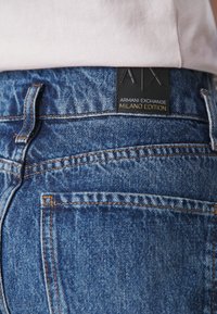 Tumši zili džinsi ar oranžiem diegiem. Iespējojams melns birka ar "Armani Exchange Milano Edition" izspiestu zeltā.