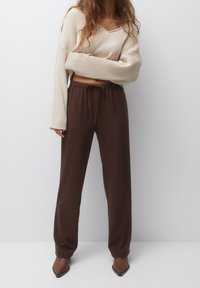 Vrouw in een crèmekleurige geribbelde gebreide cropped sweater, donkerbruine wijde broek met trekkoord en bruine puntige laarzen, staande tegen een witte muur.