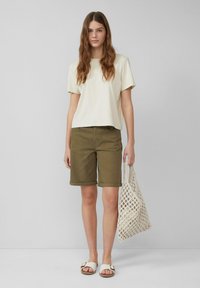 Crème t-shirt met korte mouwen gecombineerd met olijfgroene opgerolde shorts. Het model houdt een witte netten tas vast en draagt witte sandalen met een gespsluiting.