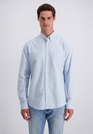 Mann trägt ein hellblaues Hemd mit Knöpfen und hellblaue Jeans, steht vor einem schlichten weißen Hintergrund.