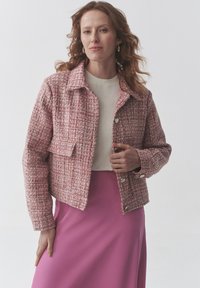 Roosa tweed jakk, millel on krae, esikott ja hõbedased nööbid. Kantakse beežist topsist üle ja sobitatakse roosa seelikuga. Sile tekstuur.