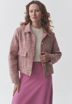 Veste en tweed rose avec un col, une poche avant et des boutons en argent. Portée sur un haut beige et associée à une jupe rose. Texture lisse.