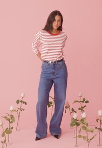 Naine seisab roosa põranda peal valgete roosidega klaasvaasides, kandes punaste triipudega särki, siniseid pükse ja musti teravate otsadega kingi, roosa tausta ees.