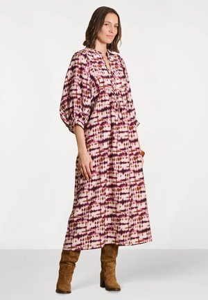 Femme portant une robe à manches longues et longueur cheville avec un motif tie-dye violet et marron, associée à des bottes en daim marron mi-mollet.