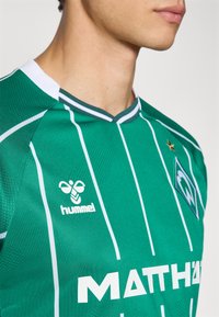 Green sports jersey with white vertical stripes, featuring a round white collar. Brand logo and lettering in white on chest. Textured fabric.