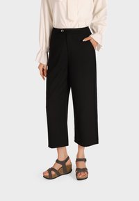 Pantaloni neri a culotte con taglio dritto e chiusura a bottone, abbinati a sandali con zeppa marroni dotati di cinturini e suole texture.