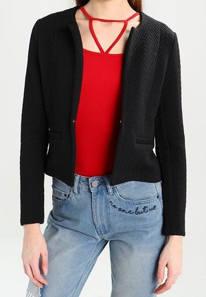 Femme portant un blazer noir texturé sur un top rouge à bretelles avec un jean bleu clair taille haute brodé "personne d’autre que nous."