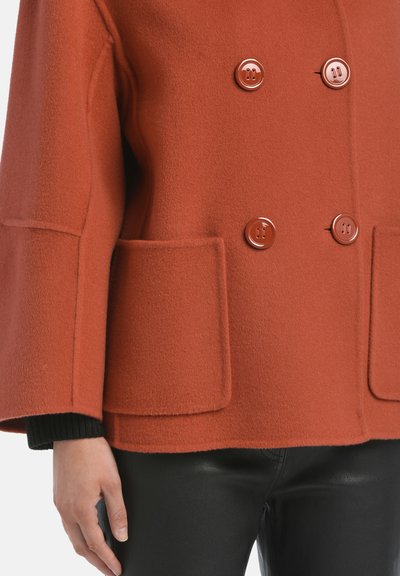 Palton din lână de culoare ruginită, cu design croi dublu, butoane maro strălucitoare și două buzunare frontale. Purtați peste pantaloni de piele negri.