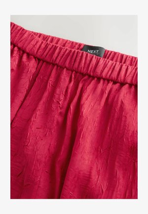 Rød krøllet stofnederdel med elastisk talje, prydet med en sort "NEXT" label øverst. Glat tekstur med flydende design.