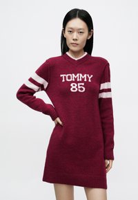 Bordó kötött ruha V-nyakkivágással, fehér "TOMMY" és "85" felirattal, csíkos ujjakkal, bordázott mandzsettákkal és szegéllyel. Simogató tapintású anyag.
