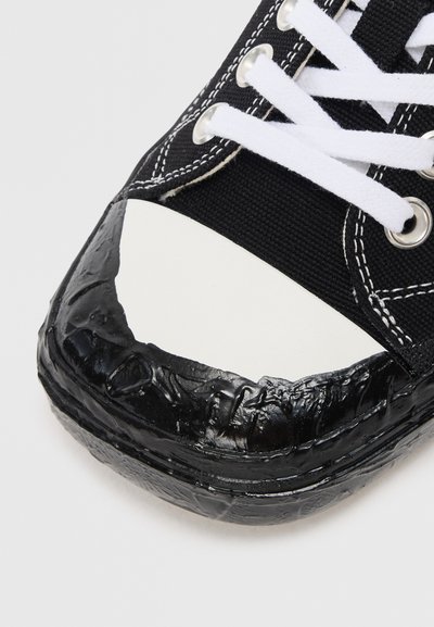 Black canvas sneaker with white rubber toe cap, black textured toe guard, and white laces. Features silver eyelets and contrasting stitching.